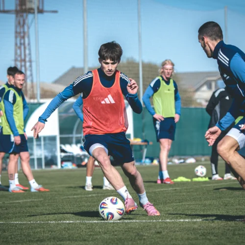 Na zdjęciu widoczna jest grupa piłkarzy trenujących na boisku, z jednym zawodnikiem w pomarańczowej koszulce, który wykonuje dribling z piłką.