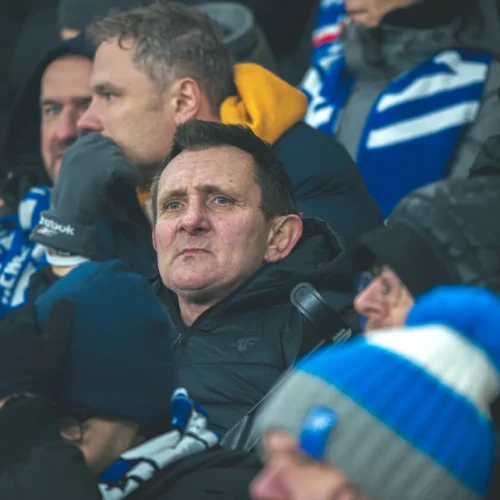 Na zdjęciu widzimy mężczyznę w czarnej kurtce, który z zmartwionym wyrazem twarzy siedzi w tłumie kibiców na stadionie, otoczony innymi osobami, które również są ubrani w zimowe odzież.