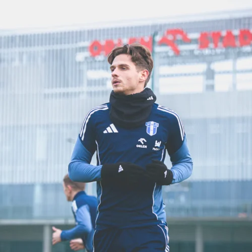 Młody piłkarz w niebieskim stroju sportowym trenuje na boisku przed nowoczesnym stadionem.