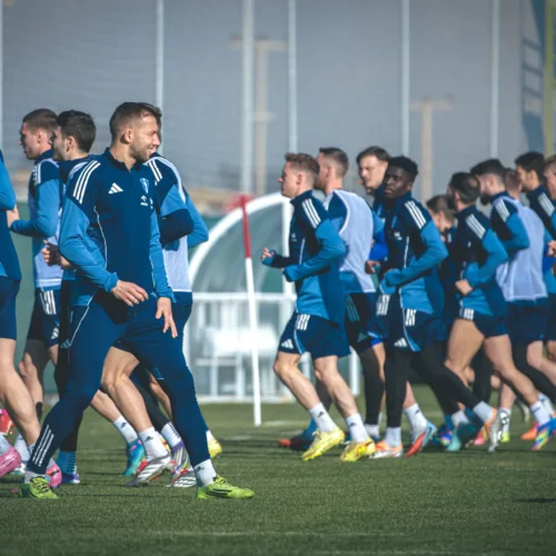 Na zdjęciu grupa piłkarzy w niebieskich strojach trenuje na boisku, biegając w słońcu.