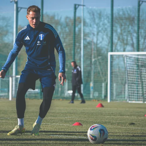 Na zdjęciu widać piłkarza trenującego na boisku, który przygotowuje się do kopnięcia piłki, w tle widać bramkę i kilka stożków treningowych.