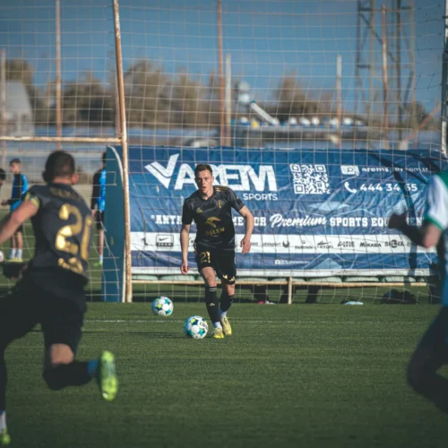 Na zdjęciu widać chwilę z meczu piłkarskiego, gdzie zawodnik w czarnej koszulce z numerem 21 biegnie z piłką na murawie, a w tle widać grupę graczy w niebieskich koszulkach oraz reklamy.