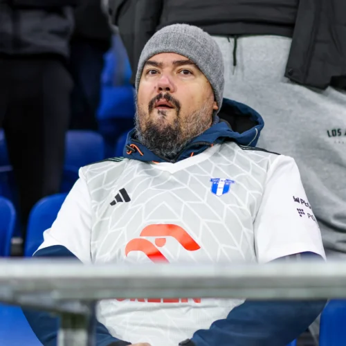 Na zdjęciu znajduje się mężczyzna w szarej czapce i białej koszulce sportowej, siedzący na trybunach w trakcie wydarzenia sportowego.