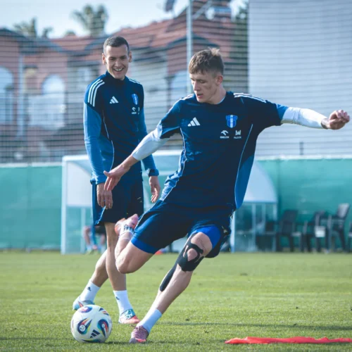 Dwaj piłkarze trenują na boisku, a jeden z nich wykonuje strzał na bramkę.