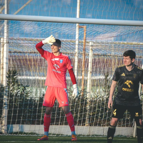 Na zdjęciu widać bramkarza w czerwonym stroju, który zasłania oczy ręką, oraz zawodnika w czarnym stroju, stojącego w pobliżu bramki na boisku piłkarskim.
