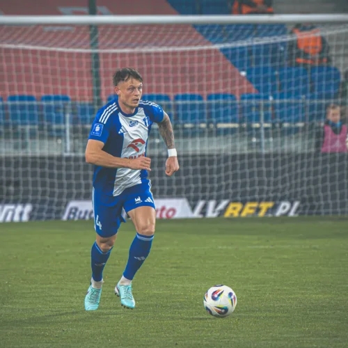 Zawodnik w niebiesko-białym stroju biega po boisku piłkarskim w kierunku piłki, z bramkarzem w tle.