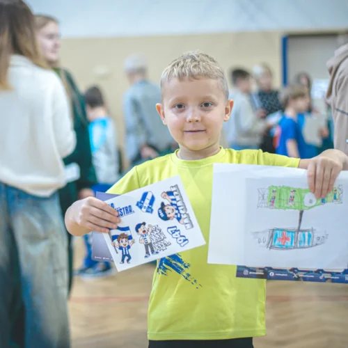 Chłopiec w żółtej koszulce trzyma rysunki i materiały promocyjne podczas wydarzenia w sali, otoczony innymi dziećmi.