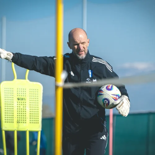 Na zdjęciu widoczny jest mężczyzna w sportowym ubraniu, trzymający piłkę nożną oraz wyciągający rękę w kierunku treningowej bramki.