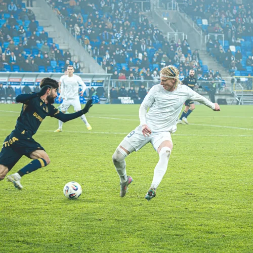 Na zdjęciu widać dwóch piłkarzy walczących o piłkę na boisku podczas meczu, w tle z sędzią i widownią.