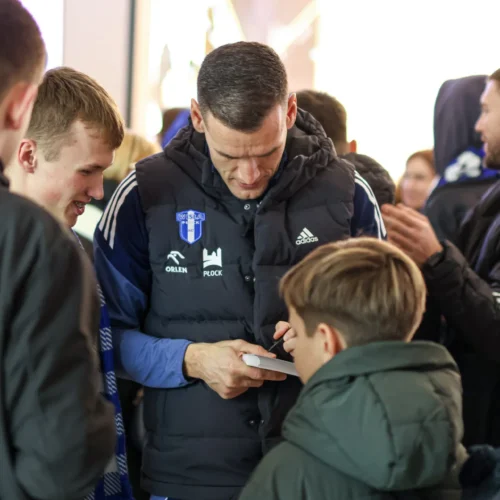 Na zdjęciu widać grupę młodych fanów, którzy z zainteresowaniem obserwują sportowca podpisującego autografy.