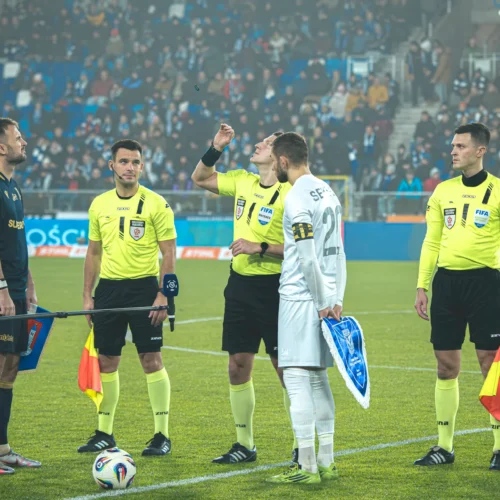 Na zdjęciu widoczni są sędziowie oraz kapitanowie drużyn podczas ceremonii przedmeczowej, przygotowujący się do rozpoczęcia spotkania piłkarskiego.