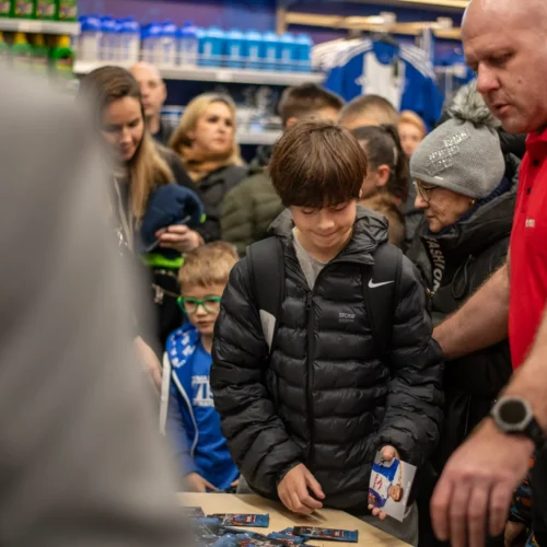 Na zdjęciu widać młodego chłopca w kurtce, który uśmiecha się, trzymając kartę z wizerunkiem, otoczonego przez tłum ludzi w sklepie.