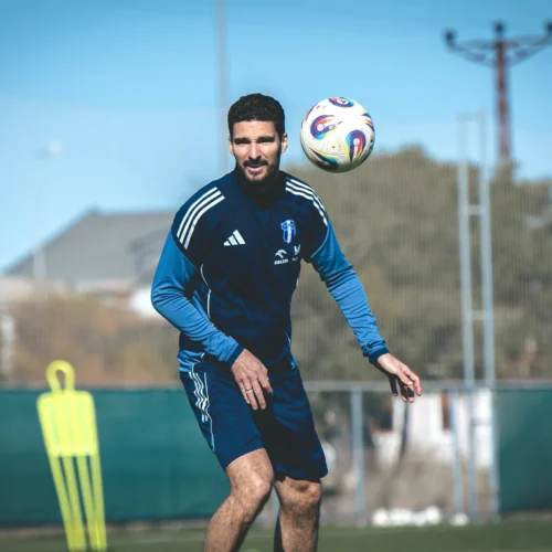 Na zdjęciu widoczny jest piłkarz w niebieskim stroju, który podnosi piłkę na boisku treningowym.