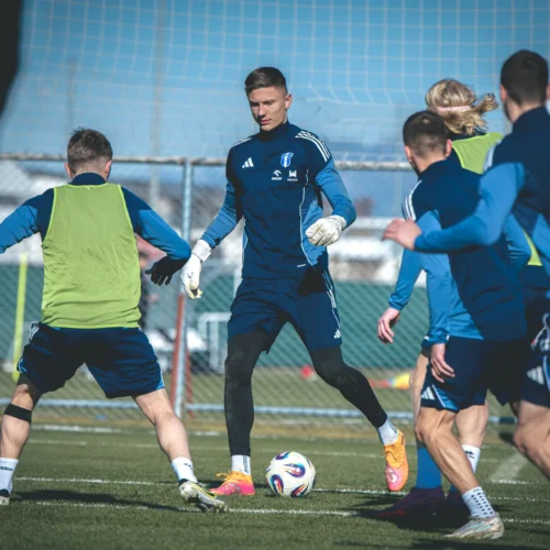 W trakcie treningu piłkarskiego grupa zawodników w niebieskich strojach ćwiczy na boisku, a w centrum uwagi znajduje się bramkarz w rękawicach.