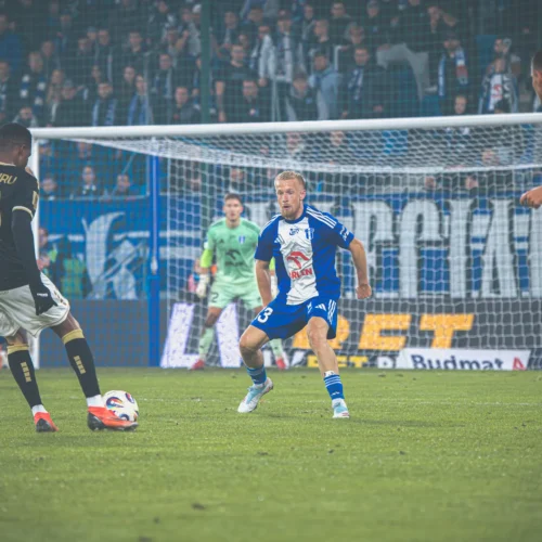 Na zdjęciu przedstawiono dynamiczną sytuację na boisku podczas meczu piłkarskiego, w którym zawodnicy w niebieskich i czarnych strojach rywalizują o piłkę.