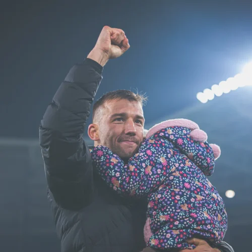 Na zdjęciu mężczyzna triumfalnie podnosi rękę, trzymając na rękach dziecko ubranego w kolorową kurtkę.