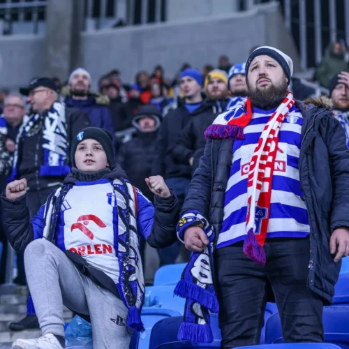 Na zdjęciu widać kibiców na stadionie, w tym dziecko i dorosłego mężczyznę, obydwaj ubrane w szaliki i stroje w barwach swojej drużyny, wyrażających emocje podczas meczu.