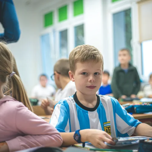 Na zdjęciu w klasie szkolnej widoczny jest chłopiec w koszulce w biało-niebieskie paski, obserwujący otoczenie, z dziećmi w tle.