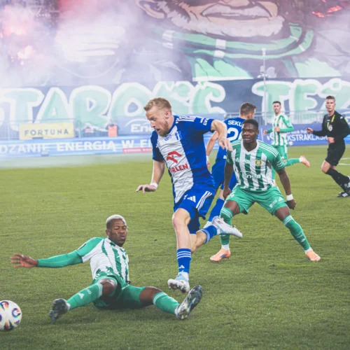 Na zdjęciu przedstawiona jest dynamiczna akcja meczu piłkarskiego, w której zawodnik w niebieskim stroju unika obrońcy leżącego na murawie, a w tle widać ognie sztuczne i transparent kibiców.