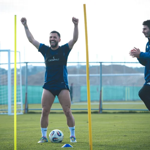 Dwóch piłkarzy cieszy się podczas treningu na boisku, jeden podnosi ręce w geście radości, a drugi klaska w dłonie.