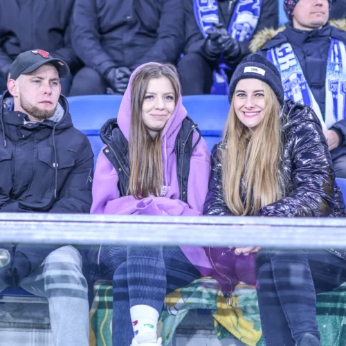 Grupa trzech młodych ludzi siedzi na trybunach, uśmiechając się i ubranych w ciepłe kurtki, podczas wydarzenia sportowego.