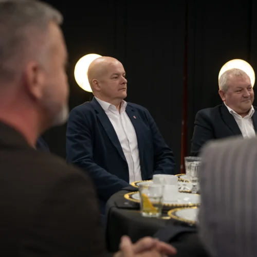 Na zdjęciu widać mężczyzn w eleganckich ubraniach, którzy siedzą przy stole podczas wydarzenia, z uśmiechami na twarzach i w tle z miękkim oświetleniem.