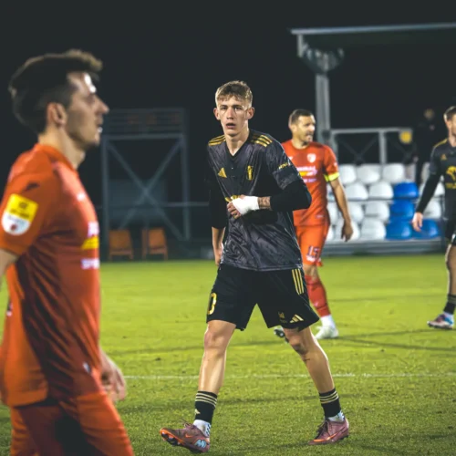 Na zdjęciu widać kilku zawodników podczas meczu piłki nożnej na murawie, a jeden z nich w czarnym stroju z widoczną kontuzją ręki wygląda na skoncentrowanego.