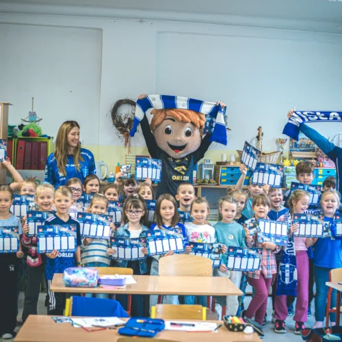 W klasie szkolnej grupa dzieci trzyma plansze z kalendarzami, a wśród nich znajduje się maskotka oraz dwie osoby dorosłe, wszyscy ubrani w niebieskie i białe kolory.