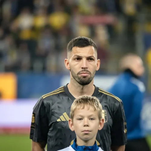 Na zdjęciu widoczny jest piłkarz w czarnej koszulce, stojący obok młodego chłopca, podczas ceremonii przedmeczowej.