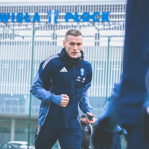 Na zdjęciu widać piłkarza trenującego na boisku w zespole Wisła Płock, w otoczeniu innych zawodników i nowoczesnego budynku w tle.