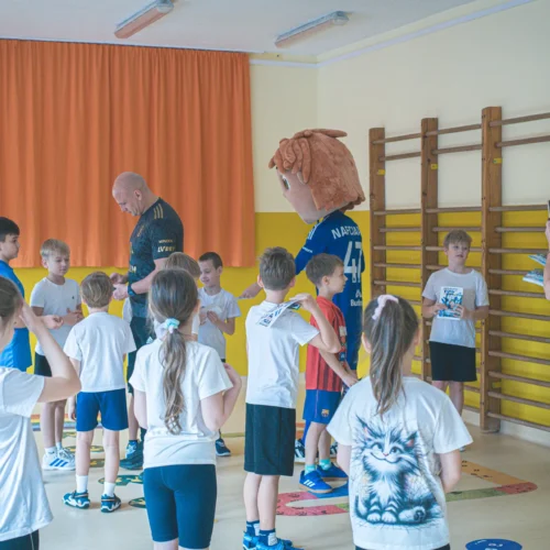 Zdjęcie przedstawia grupę dzieci w sali gimnastycznej, które są zaangażowane w zajęcia z osobą dorosłą oraz osobą w przebraniu maskotki.