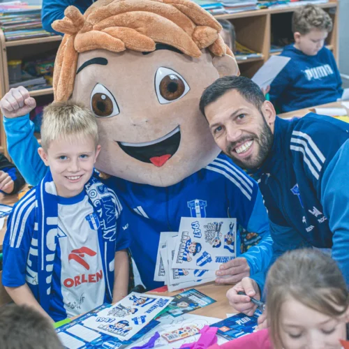 Na zdjęciu widać uśmiechniętego chłopca w niebieskiej koszulce, który siedzi przy biurku obok postaci w przebraniu maskotki, a w tle znajdują się dzieci bawiące się w klasie.