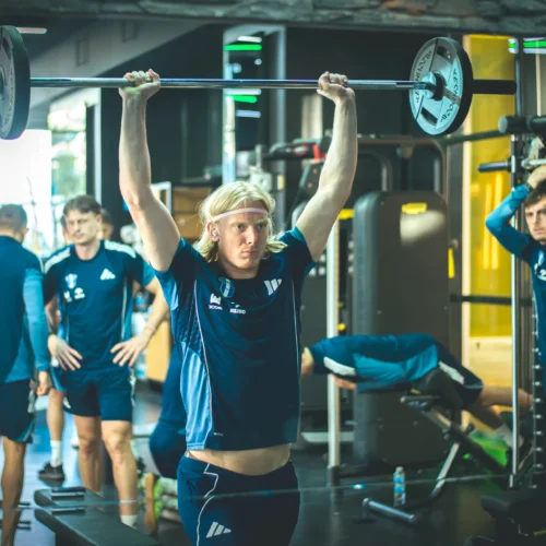 Na zdjęciu widać grupę sportowców ćwiczących w siłowni, z jednym z nich podnoszącym sztangę nad głowę.