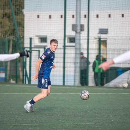 Młody piłkarz w niebieskim stroju biegnie za piłką na boisku, podczas meczu piłkarskiego.