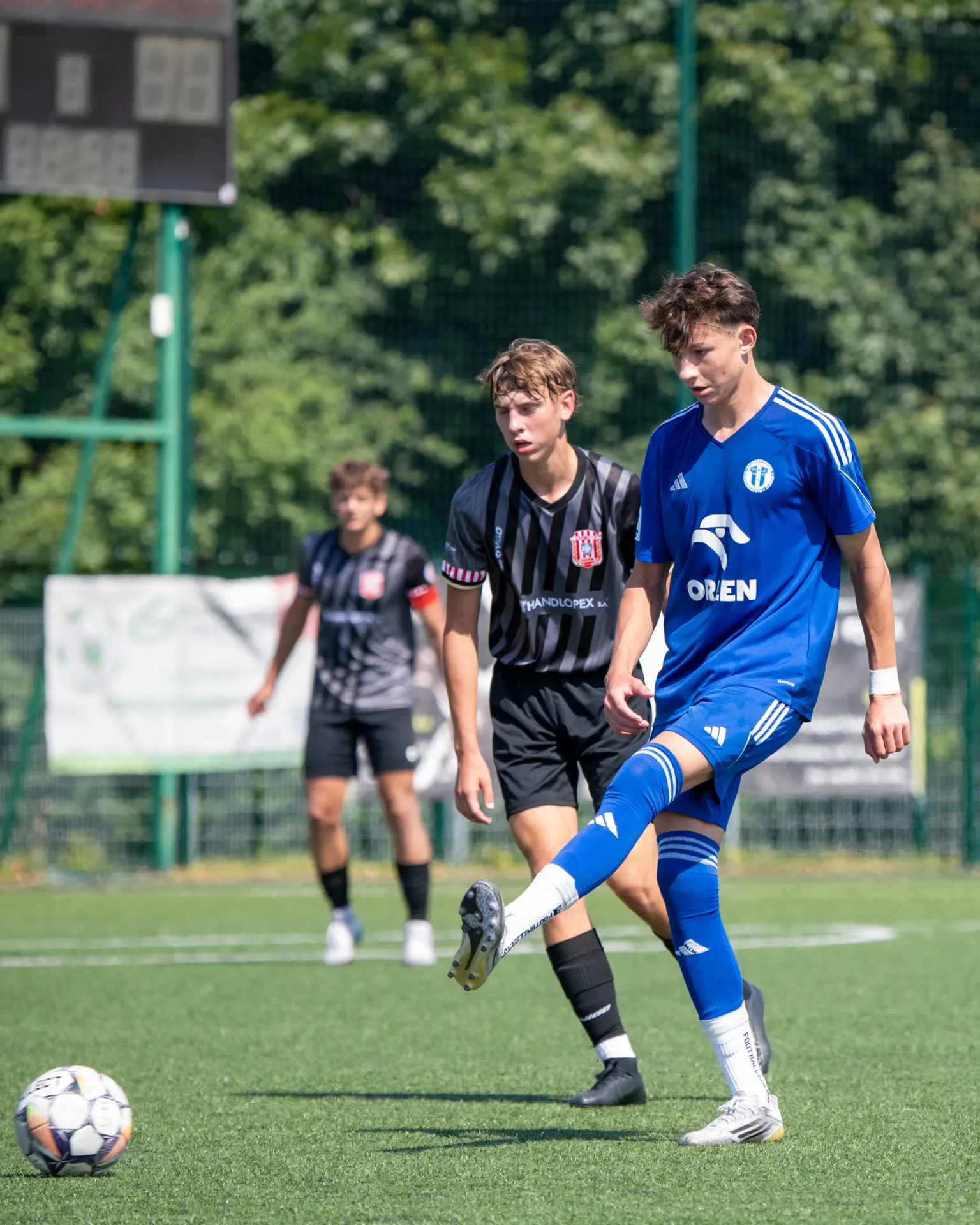Młodzi piłkarze rywalizują na boisku, a jeden z zawodników w niebieskim stroju przygotowuje się do kopnięcia piłki.