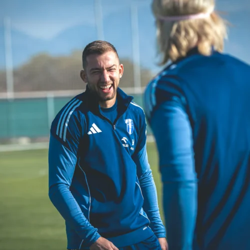 Na zdjęciu widać dwóch zawodników w niebieskich strojach piłkarskich, którzy rozmawiają i się śmieją na boisku treningowym w słoneczny dzień.