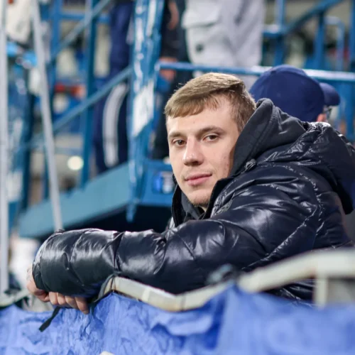 Na zdjęciu widoczny jest młody mężczyzna w czarnej kurtce puchowej, który uśmiecha się i opiera się na barierce wśród tłumu na świeżym powietrzu.