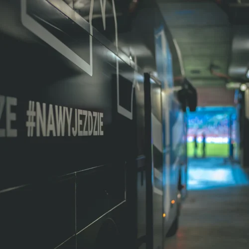 Zdjęcie przedstawia autobus sportowy stojący w tunelu, z widokiem na boisko w tle.