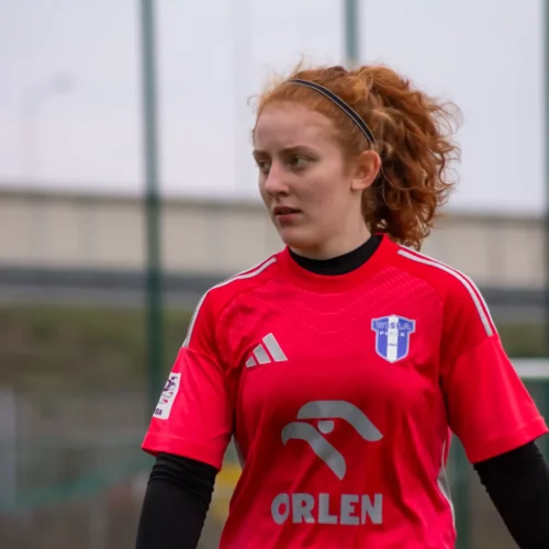 Na zdjęciu widać młodą kobietę w różowym stroju sportowym z logo drużyny piłkarskiej, stojącą na boisku.
