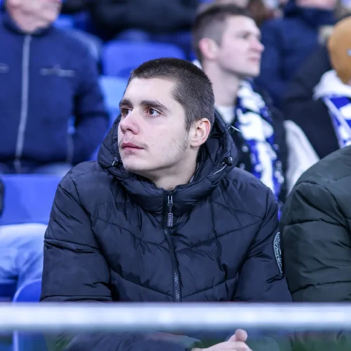 Mężczyzna w czarnej kurtce siedzi na trybunach, wpatrując się w dal podczas wydarzenia sportowego.