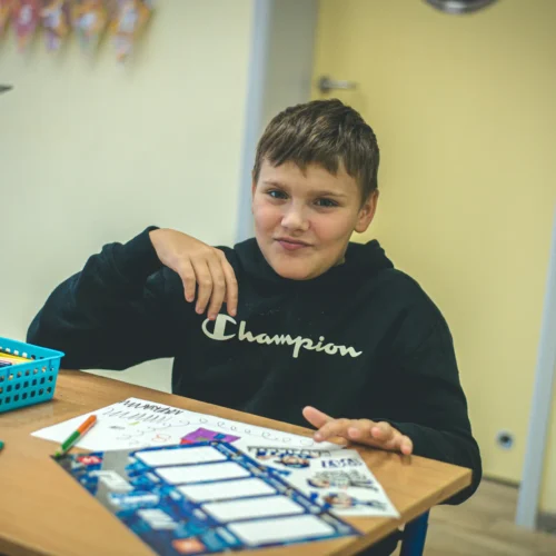 Chłopiec w czarnej bluzie siedzi przy biurku w klasie, uśmiechając się i pokazując dłonią w kierunku kamery, wokół niego znajdują się przybory do rysowania.