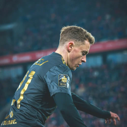 Na zdjęciu widać piłkarza w ciemnej koszulce, skupionego na grze, na tle rozmytego tłumu na stadionie.