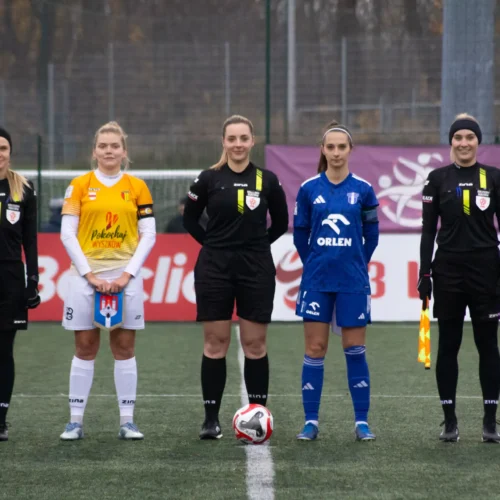 Na zdjęciu widoczna jest grupa sędziów i zawodniczek na boisku piłkarskim, w strojach reprezentacyjnych, przygotowujących się do meczu.