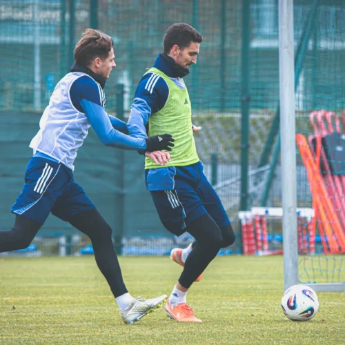 Dwaj piłkarze w trakcie treningu na boisku, jeden z nich wykonuje ruch w kierunku drugiego, który przystępuje do strzału na bramkę.