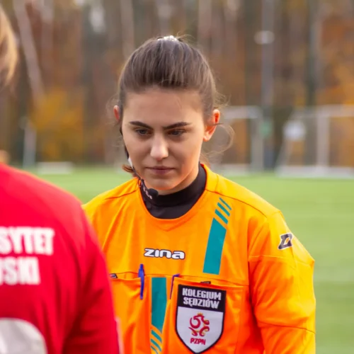 Na zdjęciu widać sędzinę na boisku piłkarskim, która w skupieniu obserwuje zawodniczkę w czerwonym stroju.