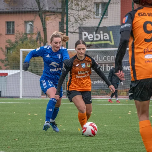 Na zdjęciu widać zawodniczki grające w piłkę nożną, z jednej strony w niebieskich strojach, a z drugiej w pomarańczowych, na boisku w deszczowej aurze.