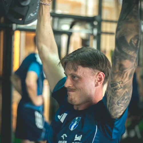 Mężczyzna w niebieskim stroju sportowym wykonuje ćwiczenie z ciężarem, podnosząc sztangę nad głowę w siłowni.