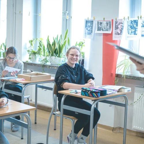 Uczniowie na lekcji w klasie, otoczeni roślinami i zdjęciami na ścianie, z uśmiechającą się dziewczyną na pierwszym planie.