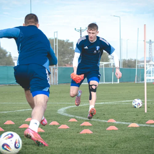 Na zdjęciu dwóch piłkarzy trenuje na boisku, przechodząc przez zestaw stożków, z jedną osobą trzymającą pomarańczowy przedmiot.