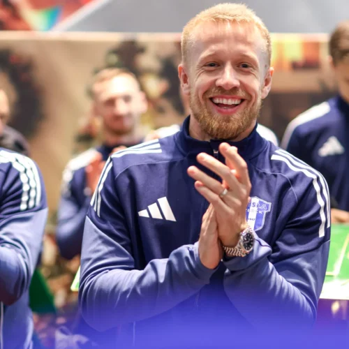 Na zdjęciu widać uśmiechniętego mężczyznę w granatowej odzieży sportowej, który klaska w dłonie w otoczeniu innych osób.